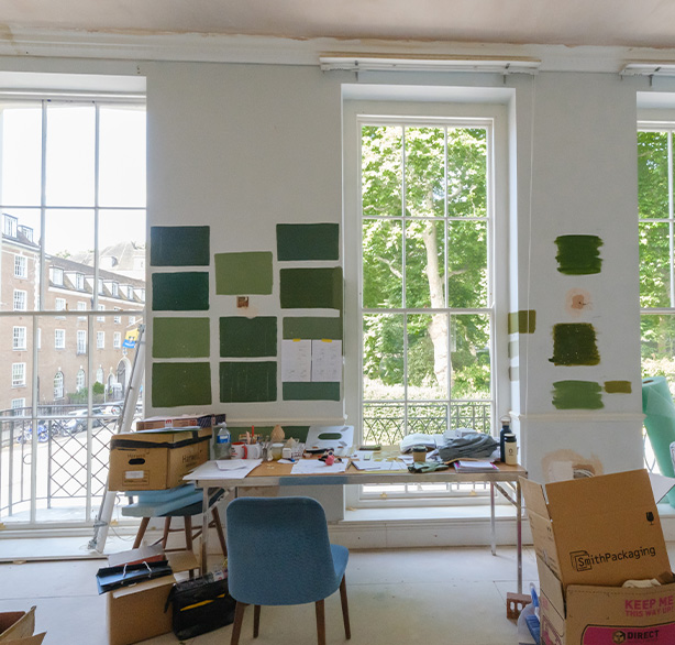 interior of an art studio with green color swatches on the walls natural light from large windows visible workspace with table and chairs creative projects in progress urban view outside the windows two shades of green paint